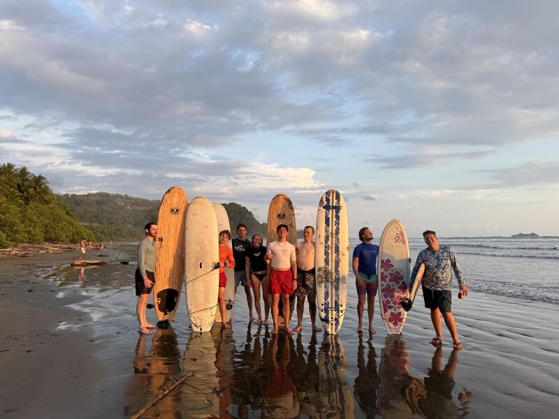 A group of about ten people stand on a wet, sandy beach with their surfboards. The surfboards are a mix of colors and sizes. The sky is cloudy with the sun peeking through, casting a warm light on the scene. In the background, there's a lush green forest and the ocean stretching out to the horizon.